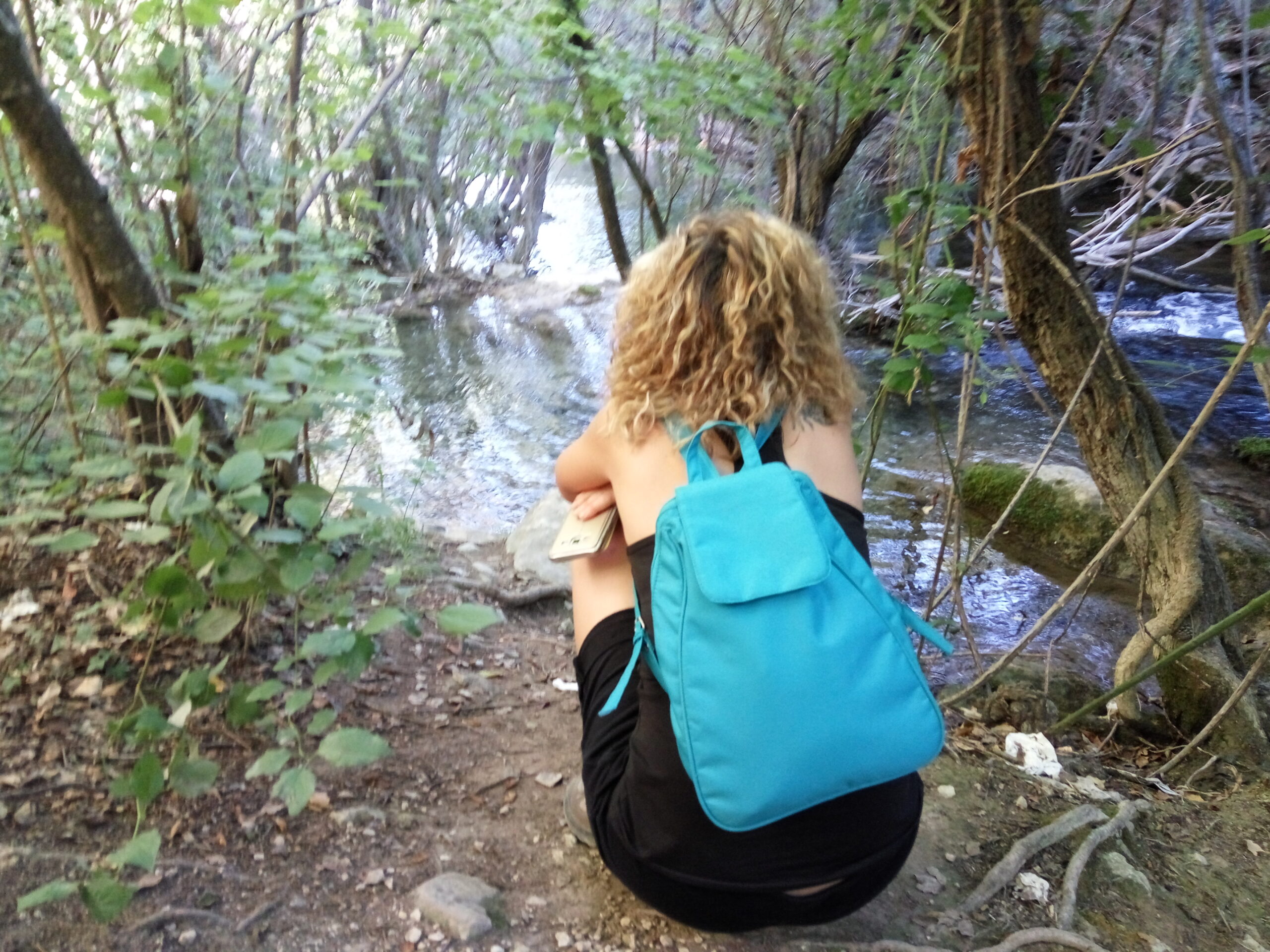 Berta caminando con mochila turquesa por un sendero junto a un río en un bosque